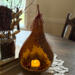 A rustic table centerpiece showing a natural gourd lit with a candle. It sits on a burlap cloth with a bouquet of twigs near.